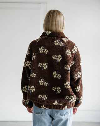 A person viewed from behind wearing a brown sherpa fleece jacket with a cream and light blue floral pattern, paired with light blue jeans, against a plain white wall.