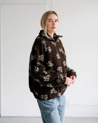 A woman wearing a brown sherpa fleece pullover with a cream floral pattern, paired with light blue jeans, standing against a plain white wall.