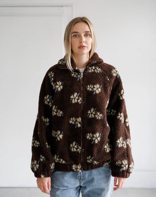 A woman wearing a brown sherpa fleece zip-up jacket with a cream and blue floral pattern, paired with light blue jeans, standing against a plain white wall.