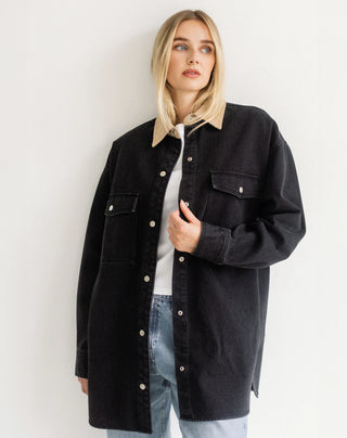 A blonde woman wearing an oversized black denim shirt jacket with a beige corduroy collar, layered over a white t-shirt and light blue jeans, standing against a plain white wall.