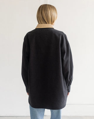 A person viewed from behind wearing an oversized black long-sleeve shirt with a contrasting beige collar, paired with light blue jeans, against a plain white wall.