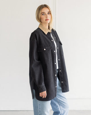 A woman modeling an oversized black denim shirt jacket with a beige corduroy collar, worn over a white top and light blue jeans against a plain white wall.