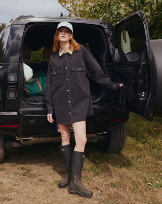 A woman stands in front of an open black SUV outdoors, wearing a dark charcoal button-front shirt dress with flap chest pockets, a light beige collar, a white baseball cap, and tall black rubber boots.