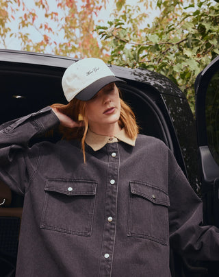 A woman wearing a dark charcoal gray denim button-up shirt with a beige collar, a white and gray baseball cap, and gold earrings, posing next to an open black car door with trees in the background.