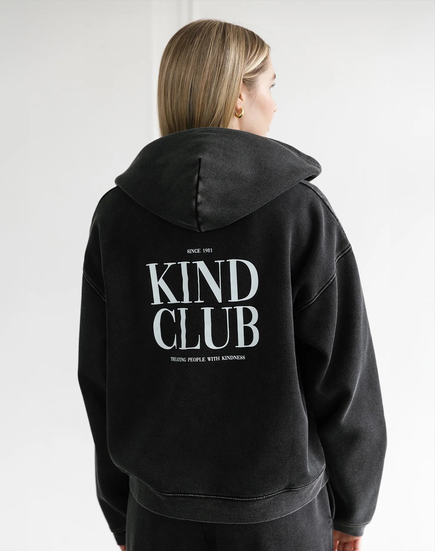 Person wearing a black hoodie with 'Kind Club' text on the back against a white background