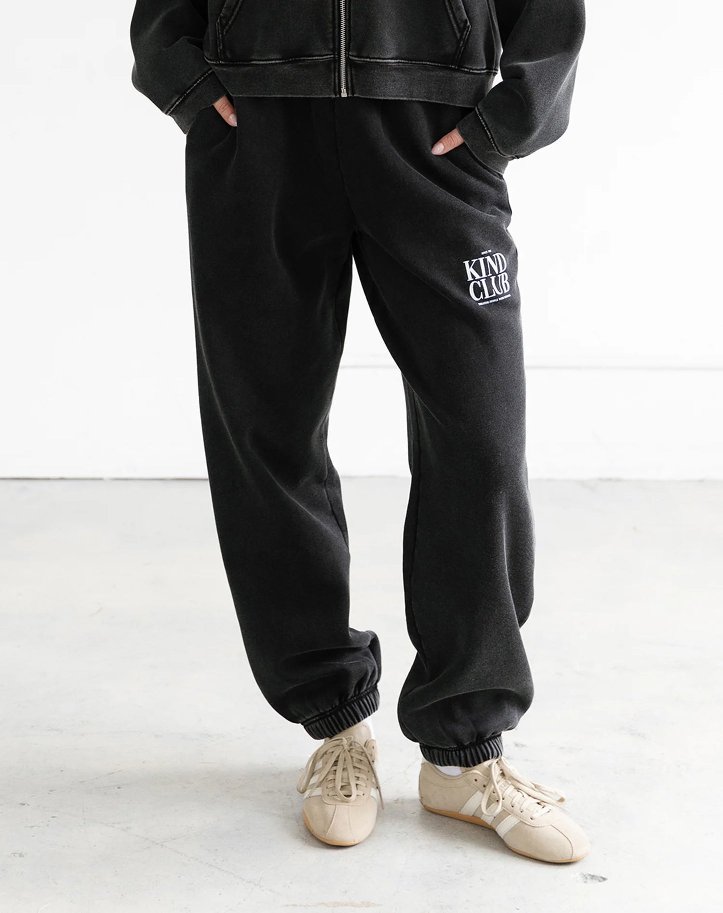 Person wearing black sweatpants with 'Kind Club' text and a black jacket on a white background