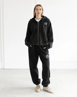 Person wearing a black hoodie and pants with a logo, standing in a white room.
