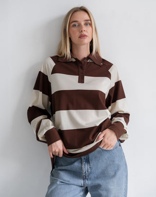 A woman wearing a brown and cream striped long-sleeve polo shirt with light blue jeans, standing against a plain light gray wall.