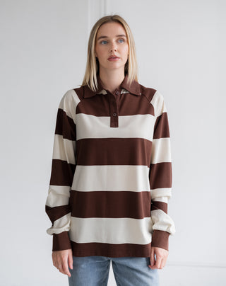 A woman wearing a brown and cream horizontally striped long-sleeve rugby shirt with a polo collar, paired with light blue jeans, standing against a plain white studio background.