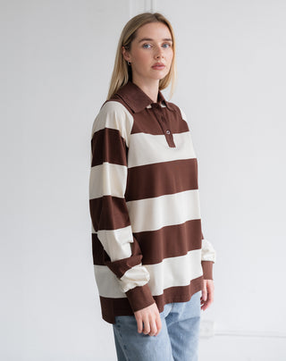 A woman modeling a brown and cream horizontally striped long-sleeve polo shirt with a buttoned collar, paired with light blue jeans, against a plain white studio background.
