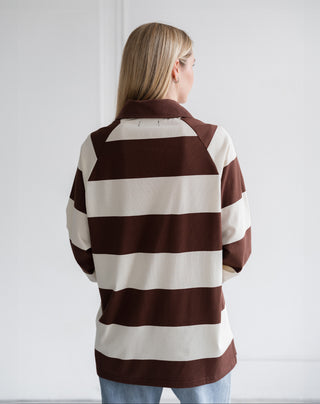 A woman viewed from behind wearing a brown and cream striped long-sleeve shirt, paired with light blue jeans, against a plain white wall.