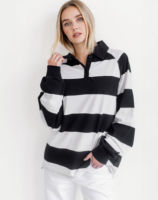 A woman wearing a black and white horizontally striped long-sleeve rugby-style shirt with a partial button placket and collar, paired with white pants, against a plain white studio background.