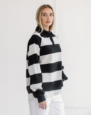 A woman modeling a black and white horizontally striped long-sleeve rugby shirt with a polo collar, paired with white pants, against a clean white background.