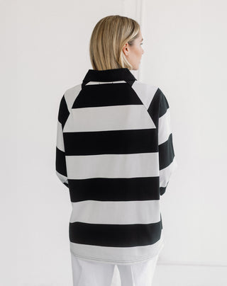 A woman shown from the back wearing a black and white horizontally striped long-sleeve top with a mock neck, paired with white pants, against a plain white studio background.