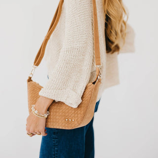 A woman wearing a cream knit sweater and blue jeans carries a tan woven leather shoulder bag with a matching strap and gold-tone hardware against a plain white background.