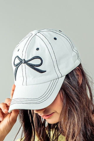 White baseball cap with a black bow design worn by a person on a gray background