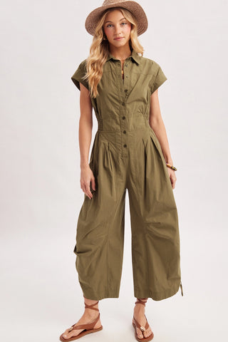 A woman modeling an olive green short-sleeve jumpsuit with a button-up front and wide legs, styled with a brown woven hat and tan sandals against a plain light gray studio background.