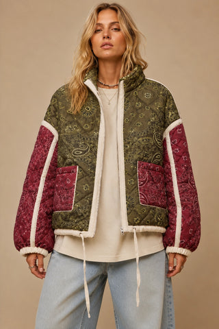 A woman wearing a patchwork quilted jacket with an olive green paisley pattern on the body and sleeves, burgundy paisley panels on the sleeves and pockets, and a cream sherpa lining, paired with a cream t-shirt and light blue jeans against a beige background.