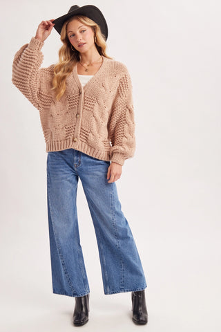 A woman modeling a chunky cable-knit cropped cardigan in a warm beige color, paired with wide-leg blue jeans, a black wide-brim hat, and black ankle boots, against a plain light gray studio background.