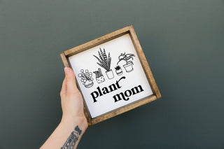 A person holding a square-shaped wooden frame sign with the text 'plant mom' and illustrations of plants.