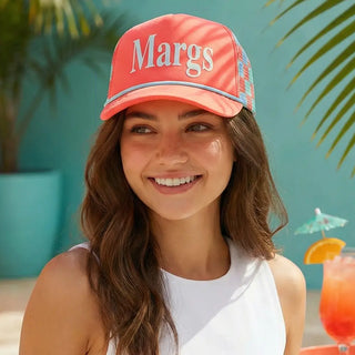 Woman wearing a pink cap with 'Margs' text, standing against a tropical background with a drink and umbrella.