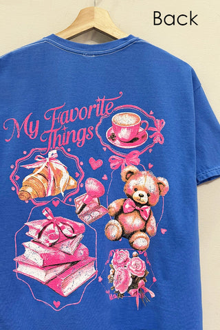 A blue short-sleeve T-shirt hanging on a wooden hanger against a light beige wall. The back of the shirt features a large pink and white graphic print with the text 'My Favorite Things' and illustrations of a croissant, a cup of coffee, books, a teddy bear, and roses.