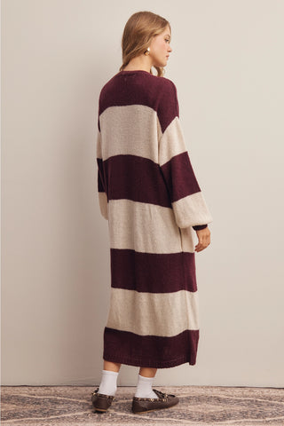 A woman wearing a long-sleeve, oversized knit sweater dress with wide horizontal stripes in burgundy and cream, shown from a side-back angle against a neutral studio background.
