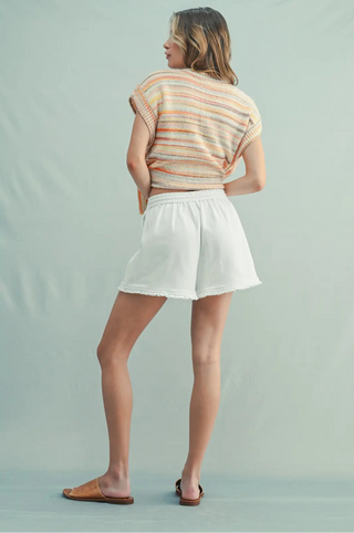 A woman is shown from the back wearing a short-sleeve knit top with thin horizontal stripes in beige, orange, and white, paired with white shorts and brown slide sandals, against a solid light blue-gray studio background.