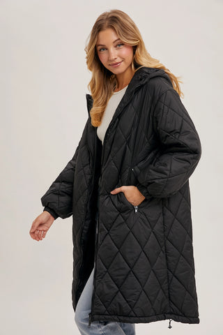 A woman modeling a long black diamond-quilted puffer coat with a hood and front zipper, paired with a white top and light blue jeans, against a plain light gray studio background.
