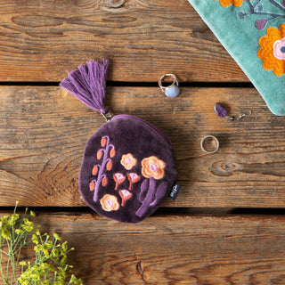 A purple velvet coin purse with a tassel and embroidered flowers, displayed on a rustic wooden surface alongside jewelry and a floral textile.