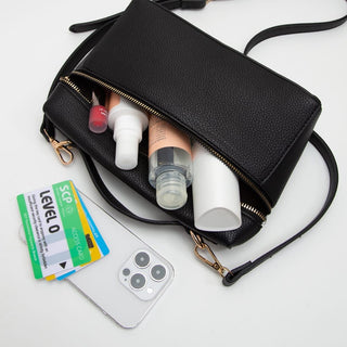 A black pebbled leather crossbody bag with gold-tone hardware, shown open with its zipper extended to reveal storage space filled with cosmetics and skincare items. A white smartphone and cards are placed beside it on a clean white background.