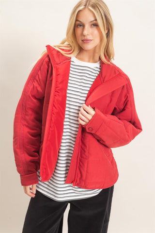 A woman wearing a bright red quilted puffer jacket over a black and white striped t-shirt and black pants, photographed against a neutral beige studio background.