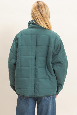 A woman viewed from behind wearing a dark teal-green quilted puffer jacket and blue jeans, standing against a plain off-white background.