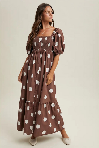 A woman modeling a brown smocked maxi dress with white polka dots, featuring puff sleeves and a tiered skirt, set against a plain beige studio background.