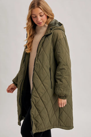 A woman wearing a long olive green quilted puffer coat over a beige ribbed turtleneck sweater and black pants, photographed against a plain light gray studio background.