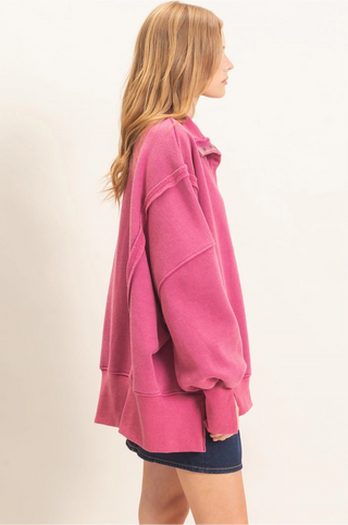 A woman shown from the side profile wearing an oversized pink sweatshirt and dark denim shorts against a plain off-white studio background.