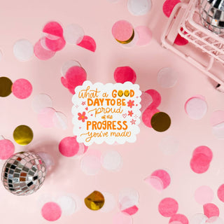 A variety of round stickers with different colors and one with a quote on it, placed on a pink surface with a pink background, alongside some metallic confetti.