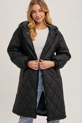 A woman wearing a long black quilted puffer coat over a white ribbed top and light blue jeans, standing against a light gray studio background.
