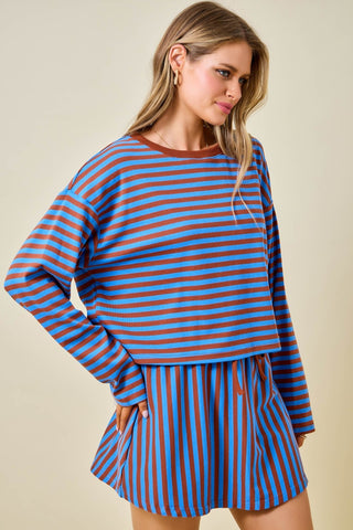 A woman modeling a matching two-piece lounge set with blue and rust-orange horizontal stripes, featuring a long-sleeve cropped top and high-waisted shorts.