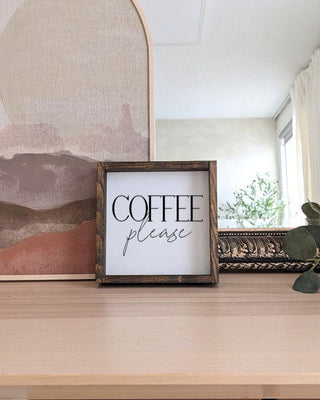 Wooden sign with 'Coffee please' text on a wooden surface with abstract art in the background.