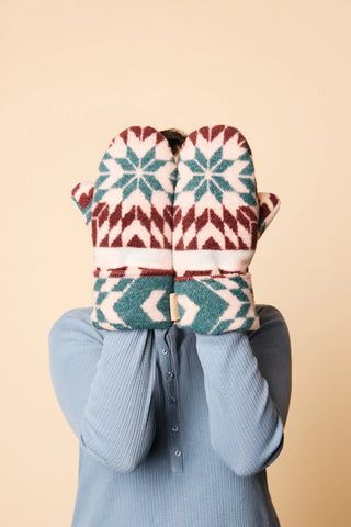 A person wearing a light blue ribbed long-sleeve top holds a pair of knit winter mittens in front of their face. The mittens have a Fair Isle pattern in teal green, burgundy red, and cream.