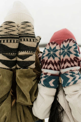 Two people bundled in winter clothing, seen from behind, wearing knit mittens with geometric and snowflake patterns. One pair is cream, black, and brown, and the other is dusty pink, teal, and white.