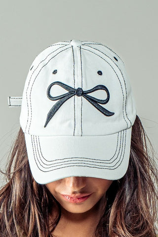 White baseball cap with a black bow design worn by a person against a neutral background