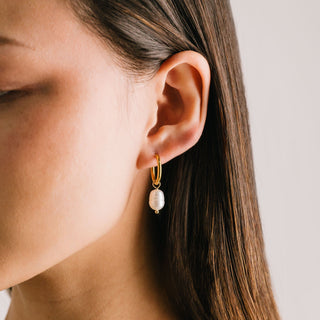 A close-up image of a woman's ear wearing a gold hoop earring with a freshwater pearl dangling below it.