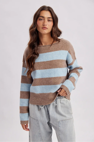 A woman wearing a knit sweater with horizontal stripes in light blue and brown, paired with light gray drawstring pants, against a plain light gray studio background.