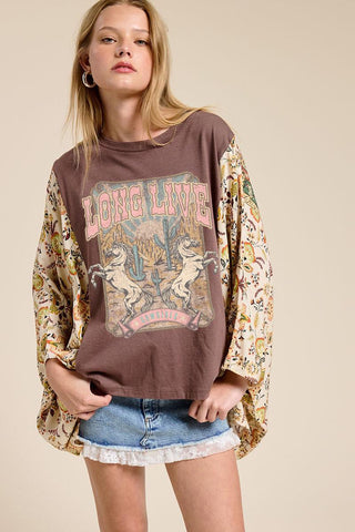 A woman modeling a brown graphic t-shirt with long, patterned bell sleeves and denim shorts against a solid beige background.