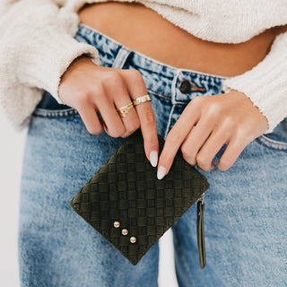 A close-up of a person holding a small black woven zip pouch with three silver studs, wearing blue jeans and a cream knit sweater.