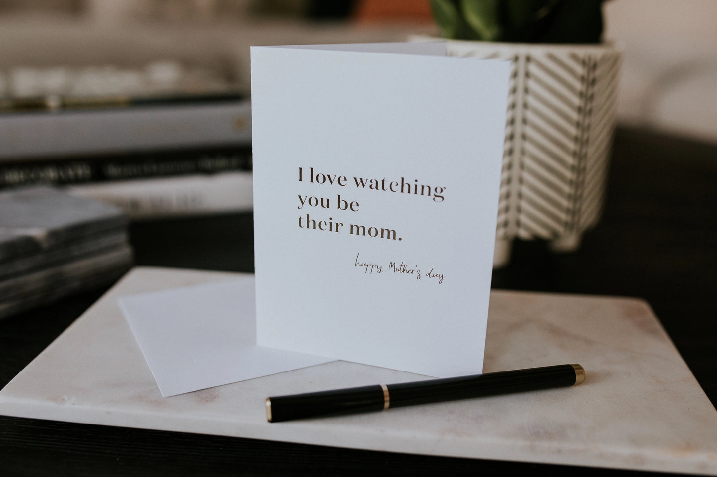 Greeting card with 'I love watching you be their mom' text on a marble surface with a pen.
