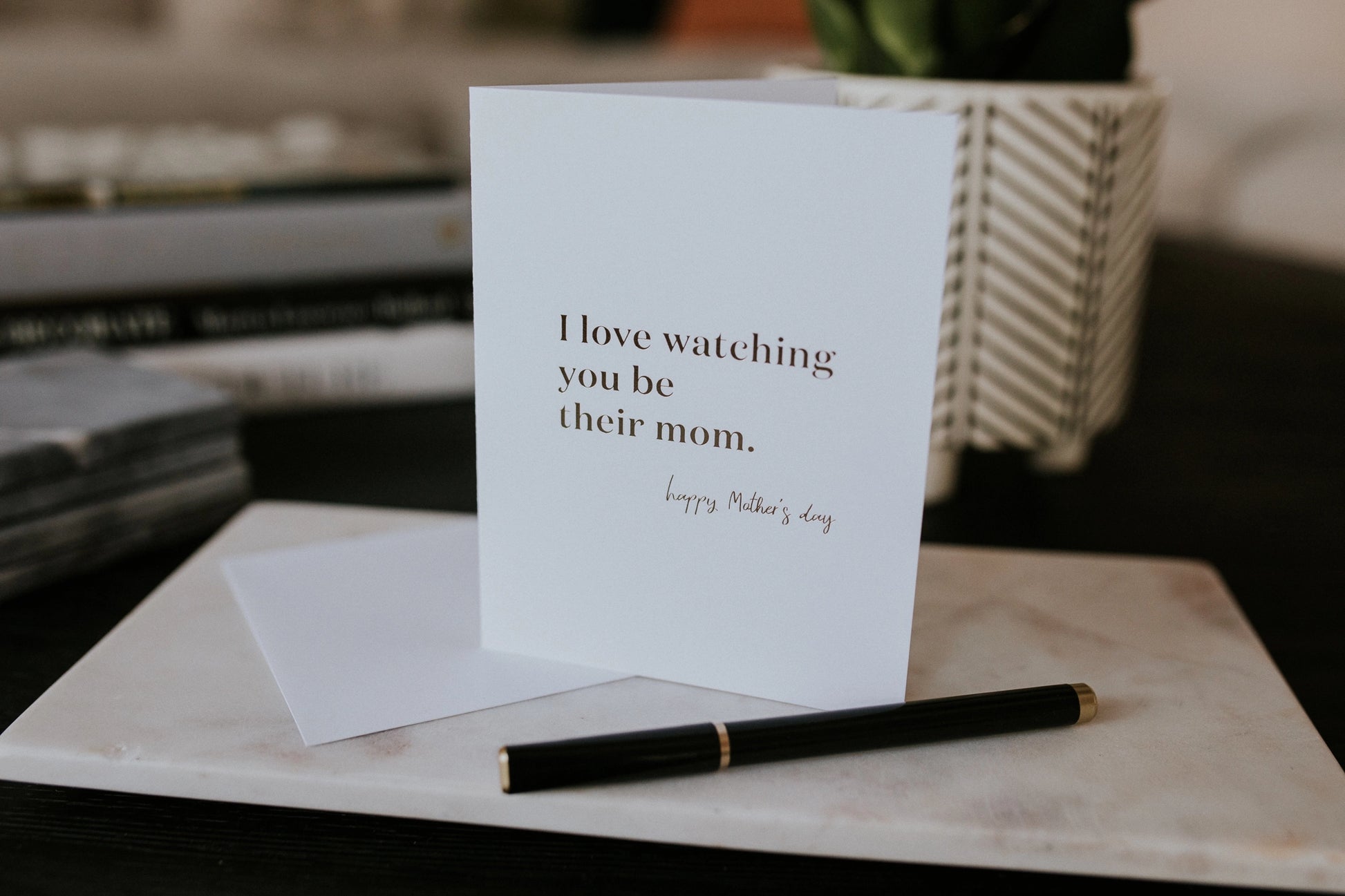 Greeting card with 'I love watching you be their mom' text on a marble surface with a pen.
