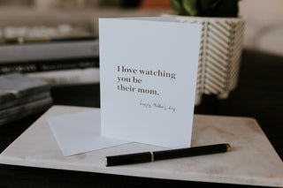 Greeting card with 'I love watching you be their mom' text on a marble surface with a pen.